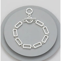A silver-toned rectangular link bracelet with a toggle clasp, displayed on a round gray stand against a light gray background.