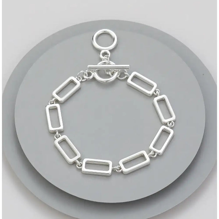 A silver-toned rectangular link bracelet with a toggle clasp, displayed on a round gray stand against a light gray background.