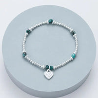 A silver beaded bracelet with small green faceted beads and a silver heart charm, displayed on a round light blue-gray pedestal.