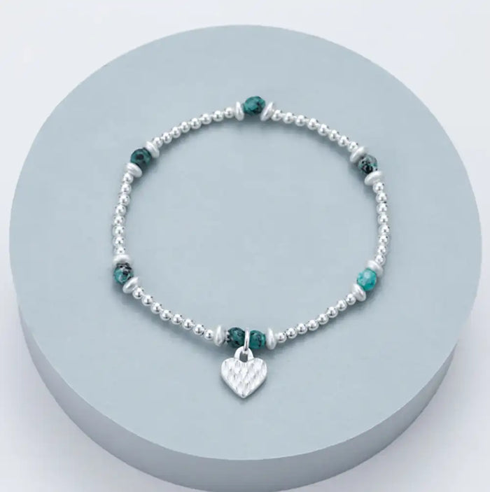 A silver beaded bracelet with small green faceted beads and a silver heart charm, displayed on a round light blue-gray pedestal.