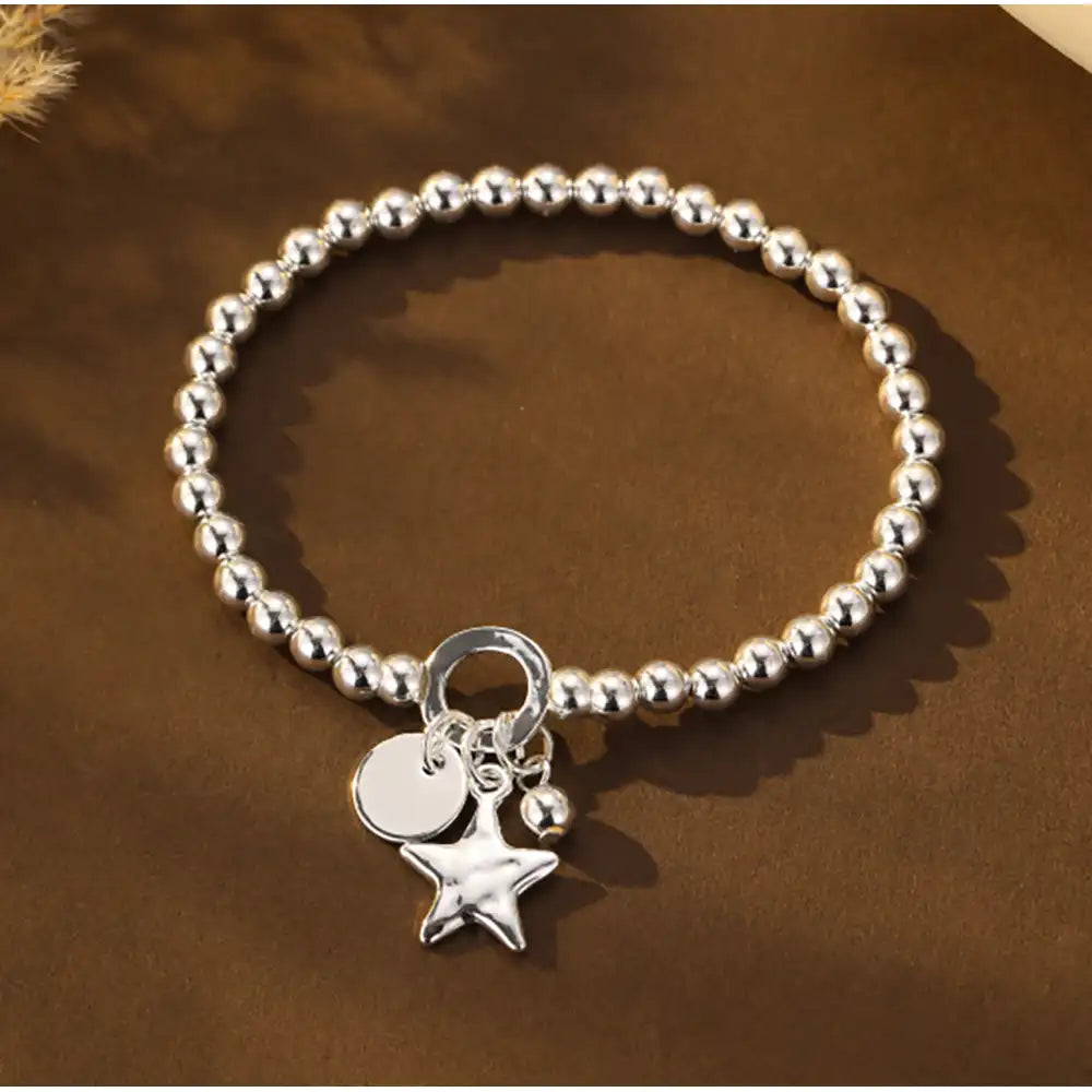 A silver beaded bracelet with a dangling star charm and two small round charms, displayed on a brown textured surface.