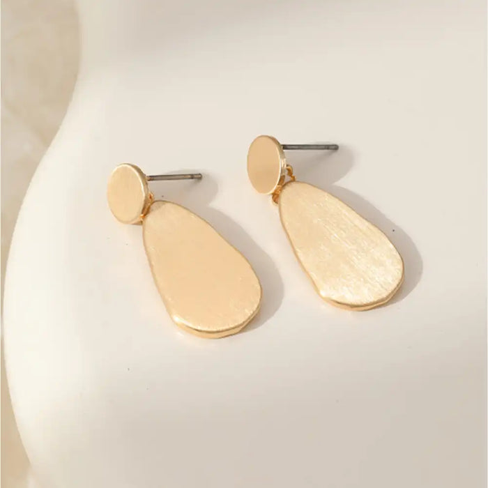 A pair of gold-toned dangle earrings with flat circular studs and irregular teardrop-shaped drops, displayed on a white curved surface.