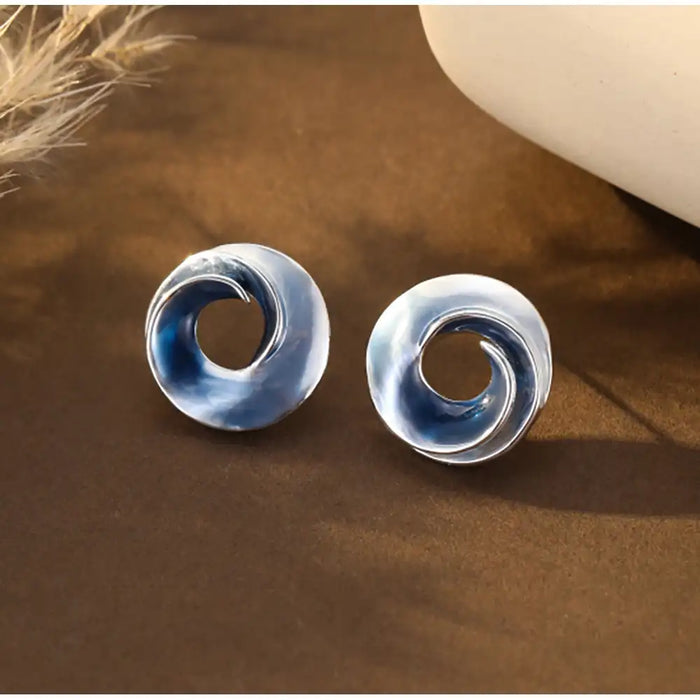 A pair of oval stud earrings with a glossy, swirled spiral design in shades of blue, gray, and silver, displayed on a dark brown textured surface.