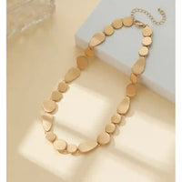 A gold-toned statement necklace made of flat, disc-like links, draped over a white geometric block on a warm beige background.