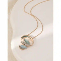 A delicate gold chain necklace with a modern abstract pendant featuring gold-tone rings, a textured light green leaf, and two small petal-shaped charms in pale blue and silver, displayed on a curved white surface.