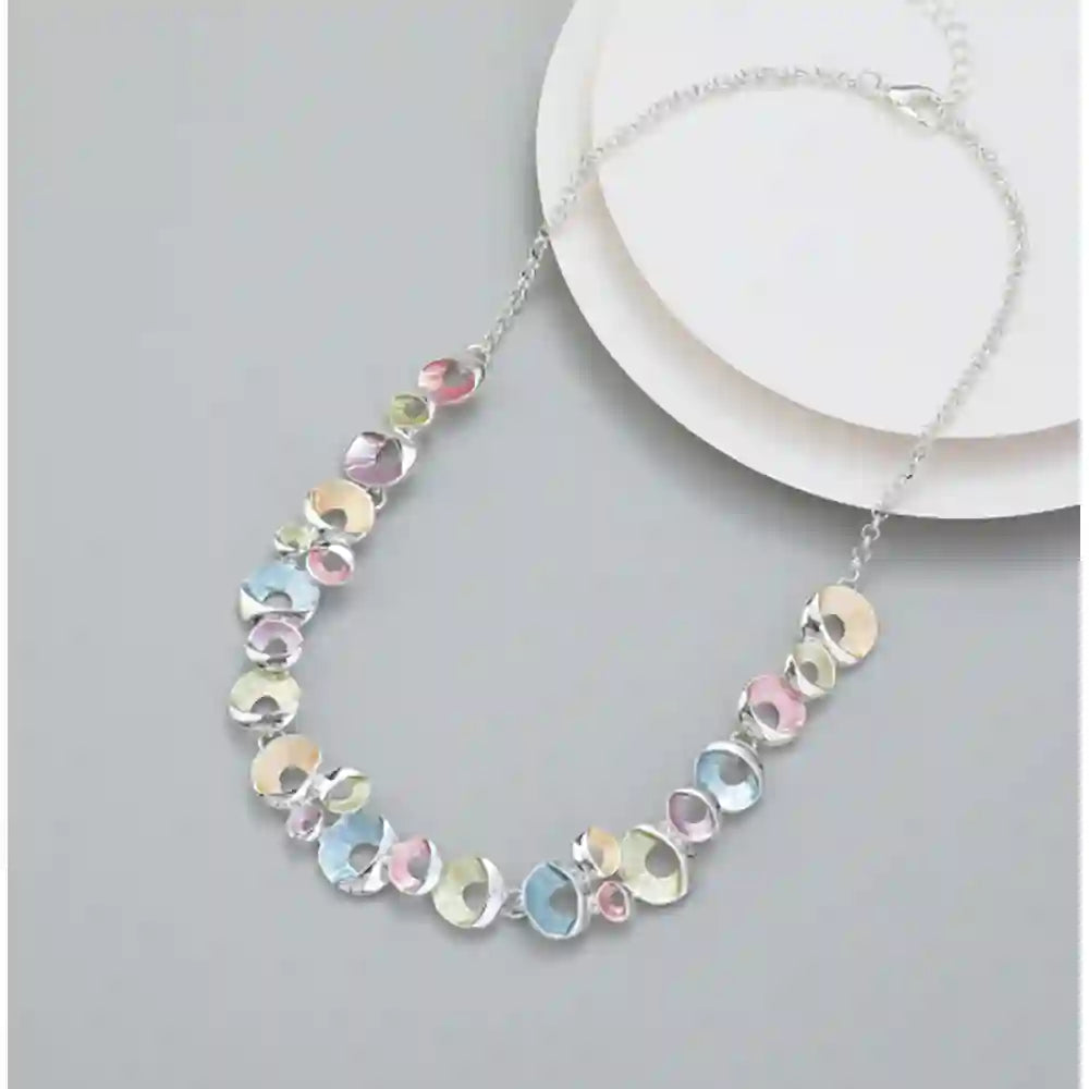 A silver chain necklace with a series of small, irregular, iridescent pastel discs in pink, blue, yellow, and green, displayed on a light gray surface next to a white plate.