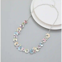 A silver chain necklace with a series of small, irregular, iridescent pastel discs in pink, blue, yellow, and green, displayed on a light gray surface next to a white plate.