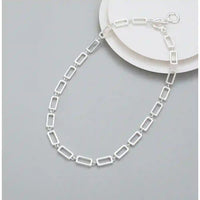 A silver rectangular link chain necklace displayed on a light gray background with a white plate prop.