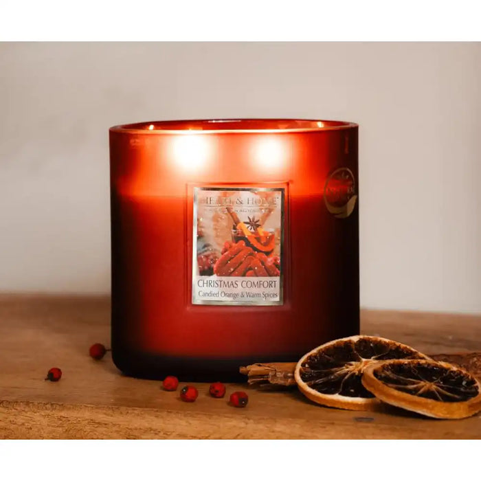 A lit red glass candle on a wooden surface with dried orange slices and small red berries beside it. 