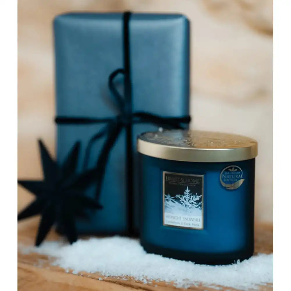 A blue glass jar candle with a gold lid sits in white faux snow in front of a matching blue wrapped gift box tied with a dark ribbon, on a wooden surface with a blurred warm background.