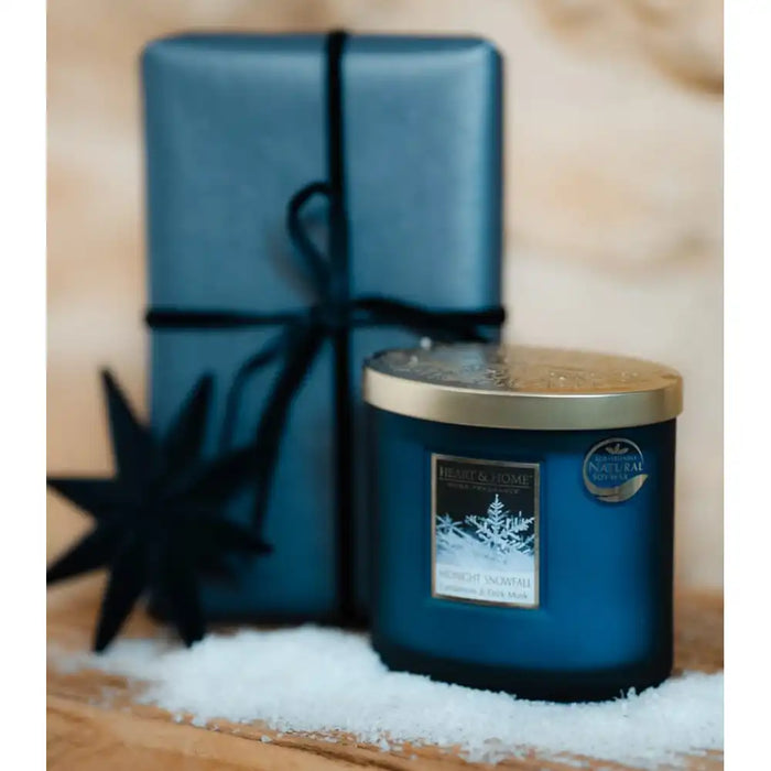A blue glass jar candle with a gold lid sits in white faux snow in front of a matching blue wrapped gift box tied with a dark ribbon, on a wooden surface with a blurred warm background.