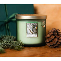 A green jar candle with a wood lid sits on a wooden surface, styled with pine needles and a pinecone. The front label reads 'Hearts & Home' and 'Shimmering Forest'.