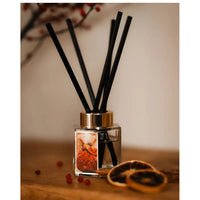 A reed diffuser with a clear glass bottle, black reeds, and a gold collar, set on a wooden surface with dried orange slices and small red berries. 