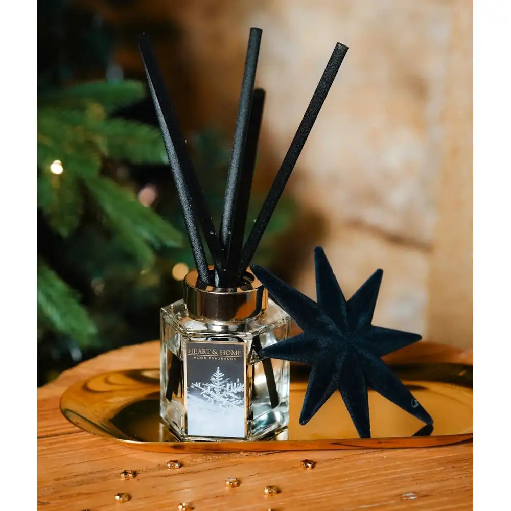 A reed diffuser bottle with black reeds and a decorative black star-shaped reed, displayed on a gold tray over a wooden surface with a blurred holiday tree in the background.