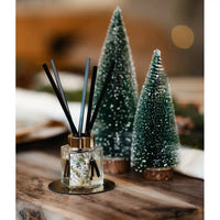 A reed diffuser with black reeds in a clear glass bottle sits on a wooden surface, styled with two small frosted Christmas tree ornaments in the background.