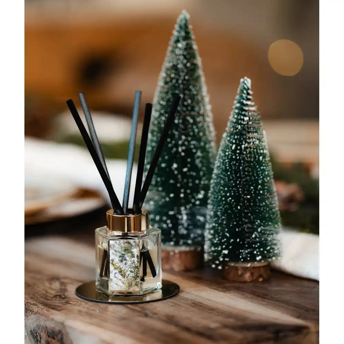 A reed diffuser with black reeds in a clear glass bottle sits on a wooden surface, styled with two small frosted Christmas tree ornaments in the background.