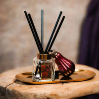 A reed diffuser with black reeds in a clear glass bottle, placed on a gold tray next to a dark red decorative bottle on a rustic wooden surface.
