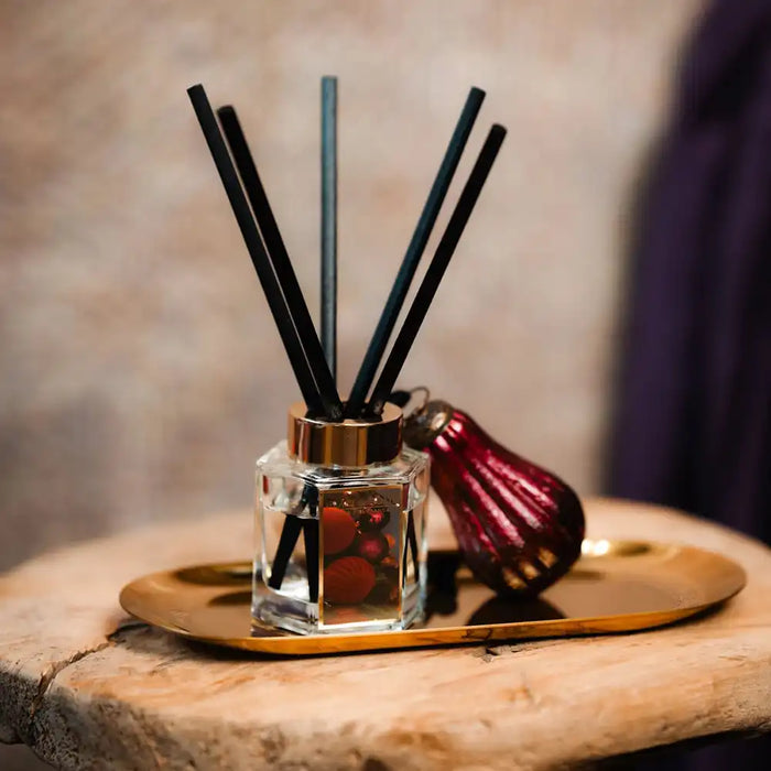 A reed diffuser with black reeds in a clear glass bottle, placed on a gold tray next to a dark red decorative bottle on a rustic wooden surface.