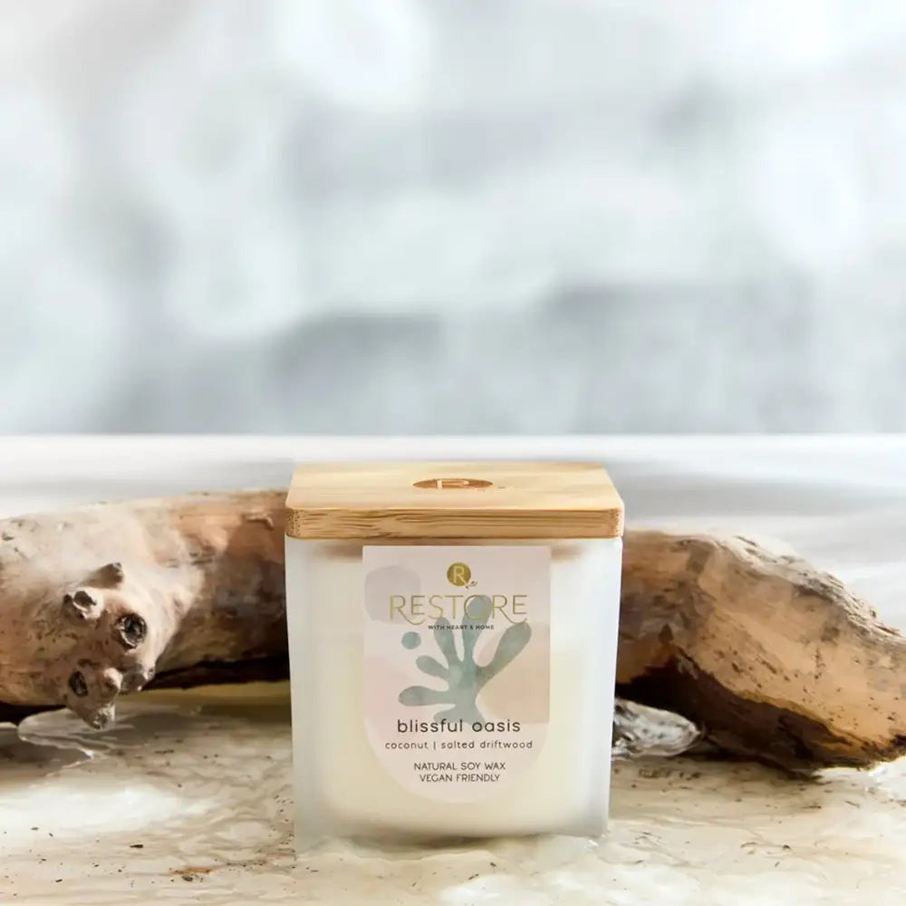 Candle labeled 'Restore' with a wooden lid on a beach setting