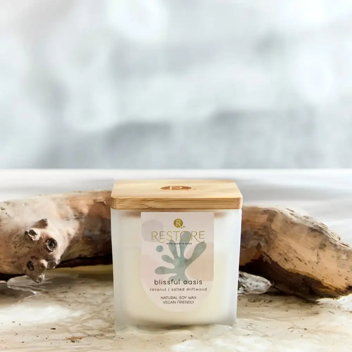 Candle labeled 'Restore' with a wooden lid on a beach setting