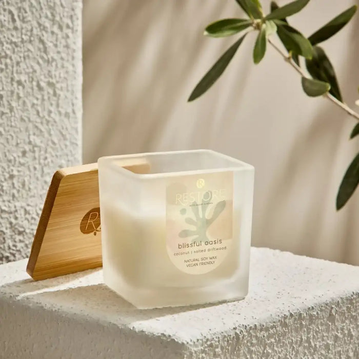 Candle labeled 'blissful oasis' with a wooden lid on a textured surface with a plant in the background.