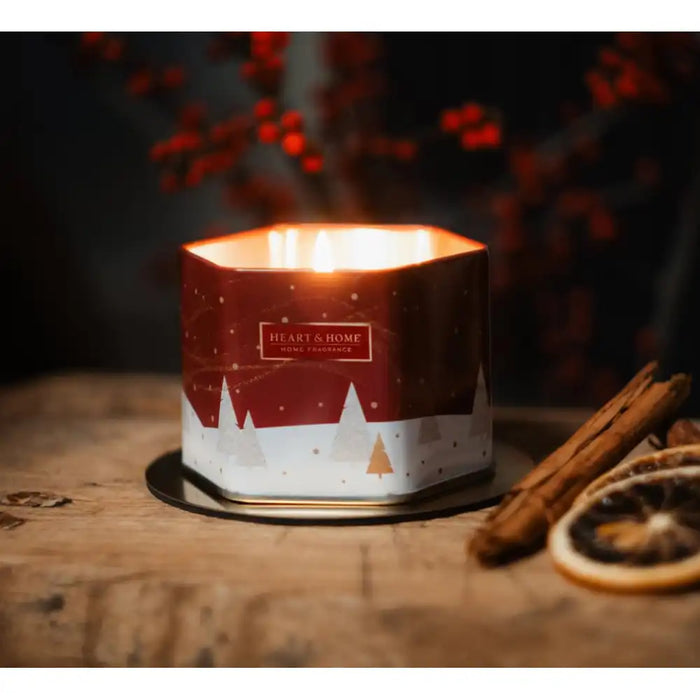 A lit hexagonal candle in a red and white tin with a winter tree design, set on a rustic wooden surface with cinnamon sticks and dried orange slices nearby.