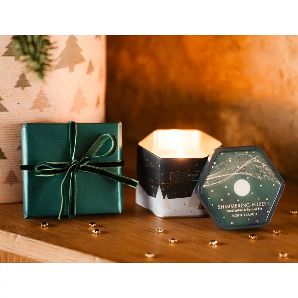 A cozy tabletop scene with a dark green wrapped gift box tied with a ribbon, a lit hexagonal candle in a white and black tin, and a hexagonal lid labeled 'Shimmering Forest' and 'Scented Candle' on a wooden surface.