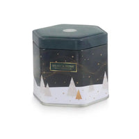 A hexagonal metal tin candle from Heart & Home, featuring a dark green lid and a winter-themed design with a dark green upper section, gold stars, and a snowy white lower section with minimalist pine trees.