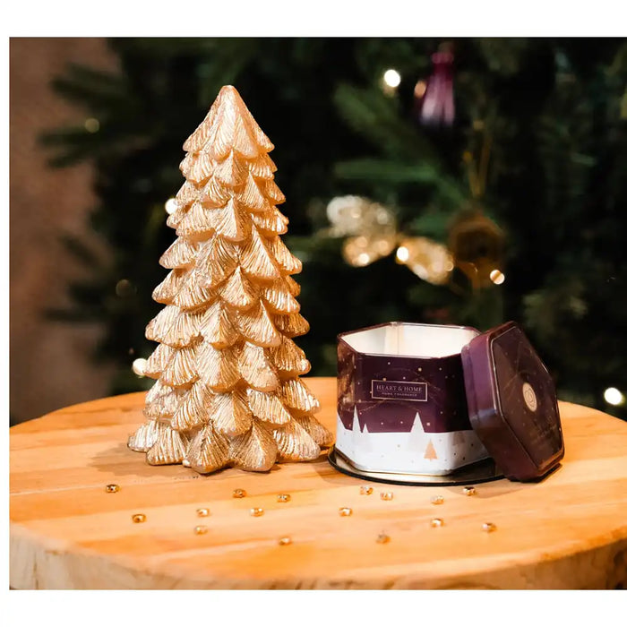 A gold textured Christmas tree ornament stands beside a purple and white tin candle on a round wooden surface, with a blurred Christmas tree in the background.