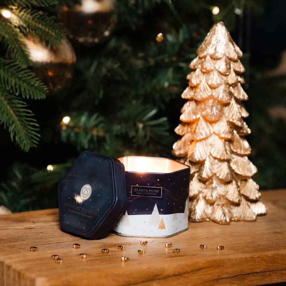 A lit hexagonal candle in a dark navy and white tin sits on a wooden surface beside a metallic gold Christmas tree ornament, with a blurred Christmas tree in the background.