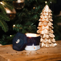 A lit hexagonal candle in a dark navy and white tin sits on a wooden surface beside a metallic gold Christmas tree ornament, with a blurred Christmas tree in the background.