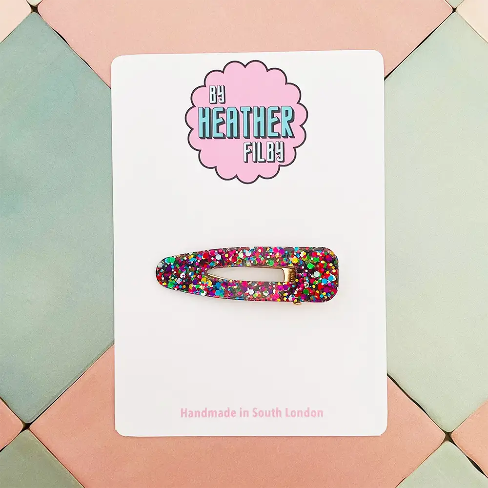Colorful glittery hair clip on a white card with 'Be Heather Filby' branding, set against a geometric patterned background.