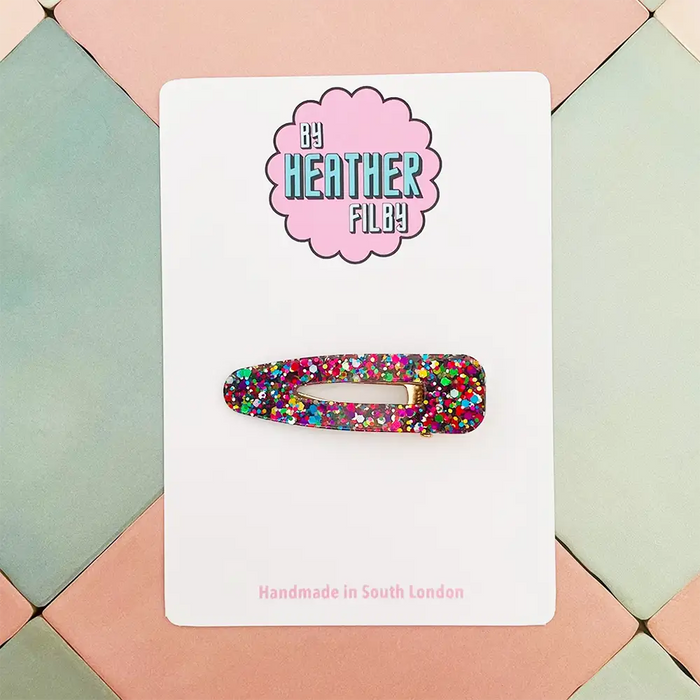 Colorful glittery hair clip on a white card with 'Be Heather Filby' branding, set against a geometric patterned background.
