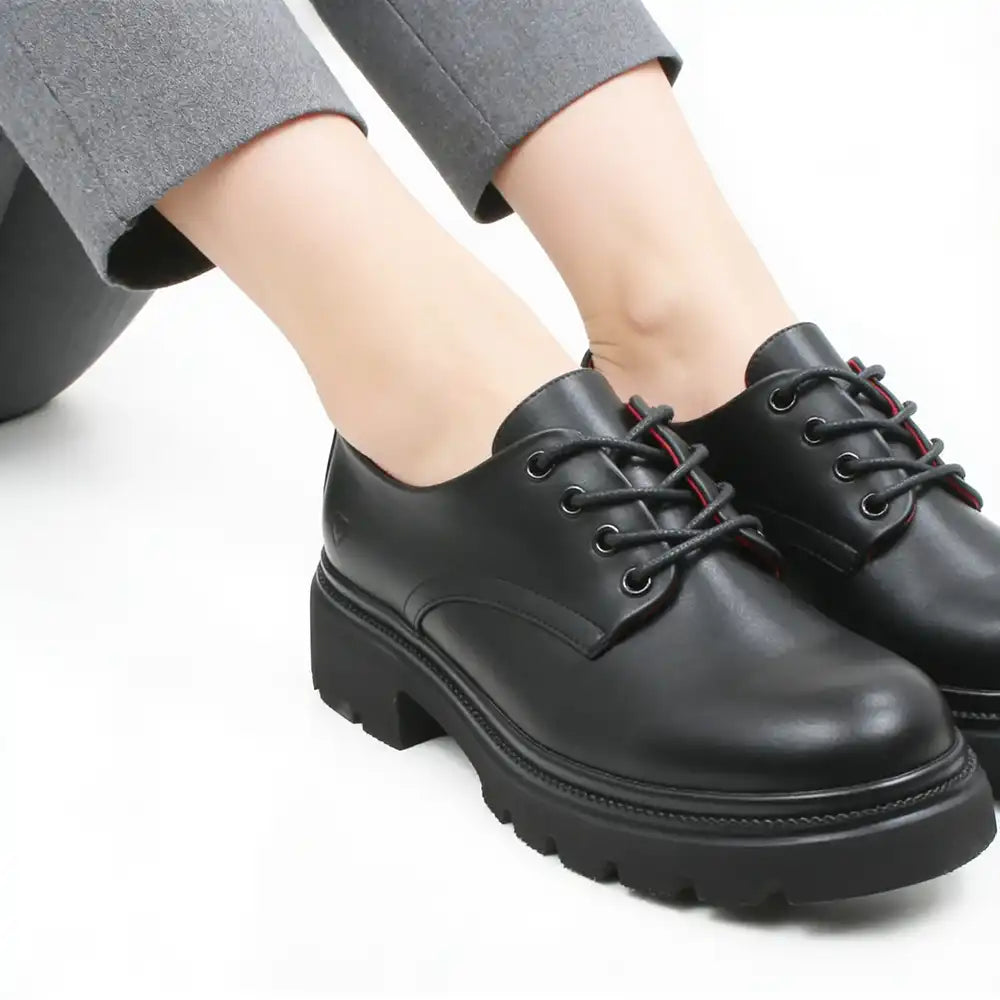 A pair of black leather lace-up oxford shoes with chunky lug soles, shown on a model wearing gray trousers against a white background.