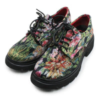 A pair of low-top lace-up shoes with a vibrant, multicolored floral and botanical print on the uppers, black laces, and thick black soles, shown against a plain white background.