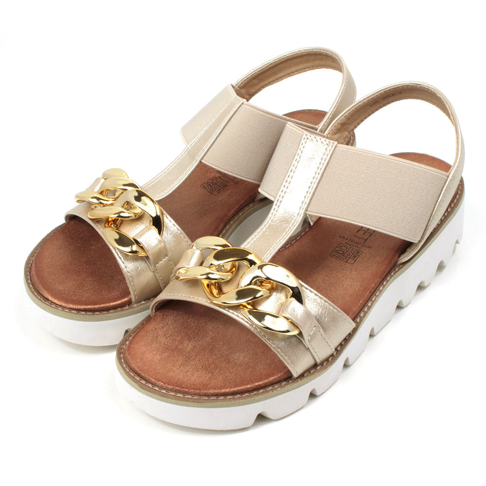 Gold low wedge sandals from Heavenly Feet. Over toes strap and upper foot elasticated strap. Strap around the back of the heel. Chunky gold chain decoration over the toes. Tan coloured footbeds. White low wedge soles with wide deep carved tread. Angled view.