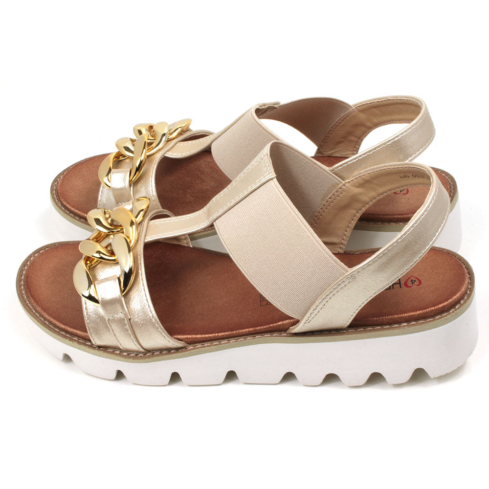 Gold low wedge sandals from Heavenly Feet. Over toes strap and upper foot elasticated strap. Strap around the back of the heel. Chunky gold chain decoration over the toes. Tan coloured footbeds. White low wedge soles with wide deep carved tread. Side view.