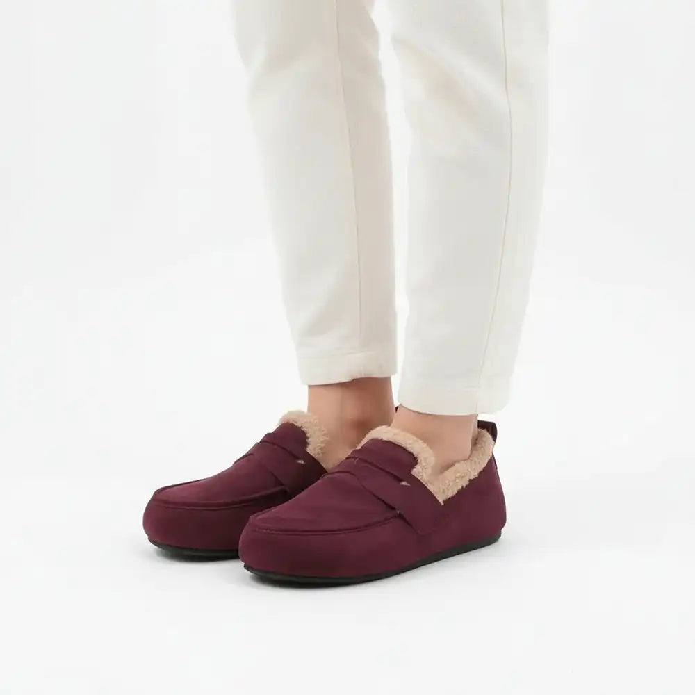A person wearing burgundy slip-on slippers with beige sherpa lining, paired with white pants, shown from the calves down against a clean white background.