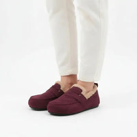 A person wearing burgundy slip-on slippers with beige sherpa lining, paired with white pants, shown from the calves down against a clean white background.
