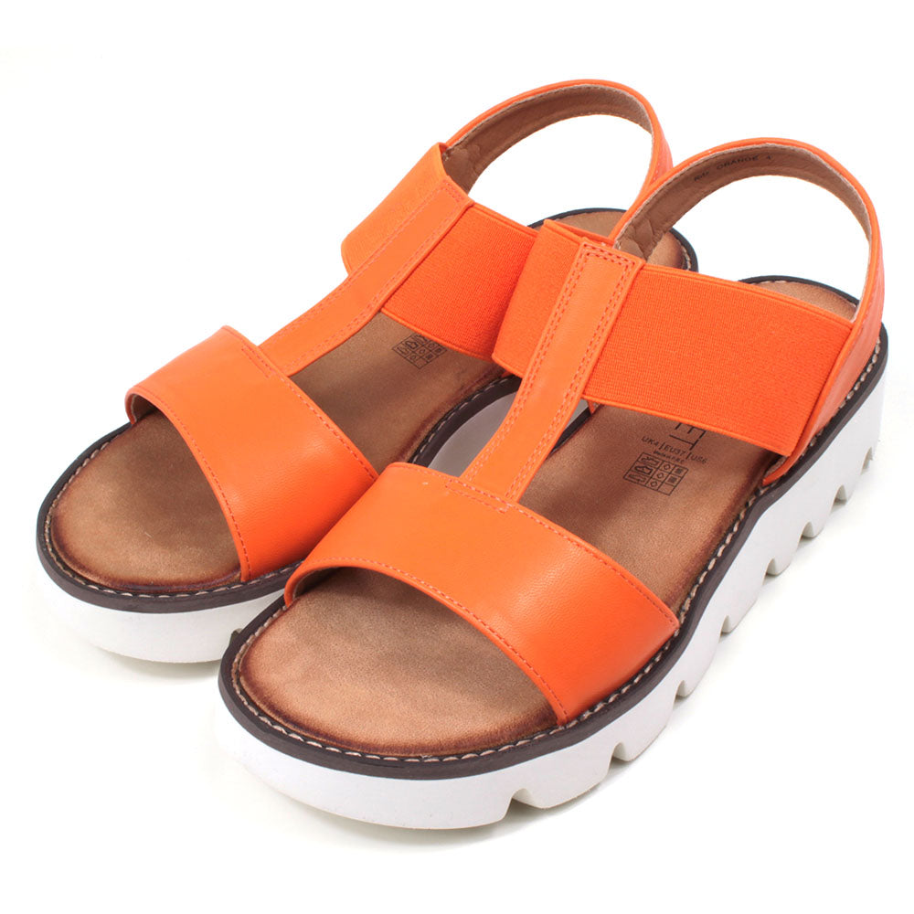 Orange low wedge sandals from Heavenly Feet. Over toes strap and upper foot elasticated strap. Strap around the back of the heel. Tan coloured footbeds. White low wedge soles with wide deep carved tread. Angled view.