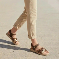 A person wearing brown and beige strappy sandals with rolled-up beige pants, walking on a light gray paved surface.