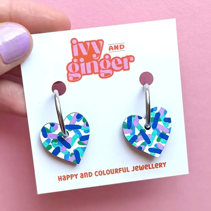 Colorful heart-shaped earrings on a 'Ivy and Ginger' branded card against a pink background.