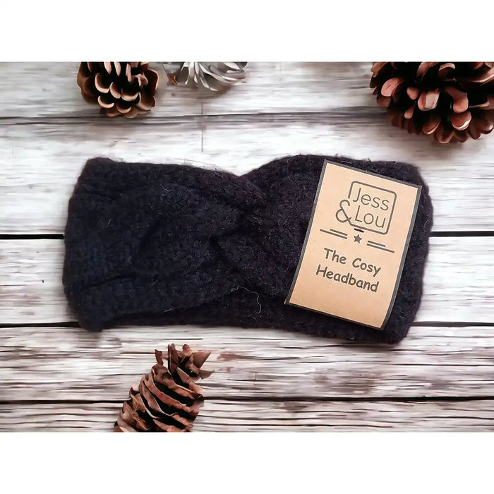 A black chunky knit headband with a front knotted detail, displayed on a wooden surface with pine cones scattered around.