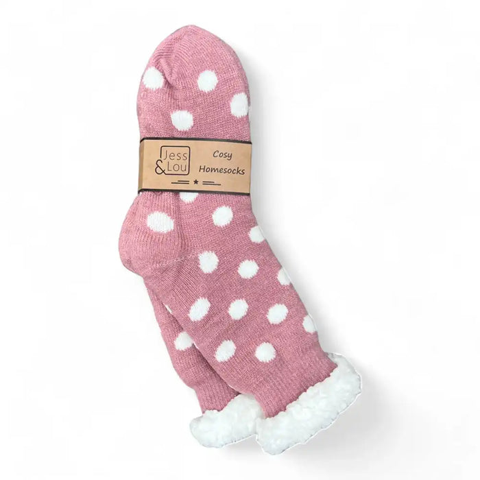 A pair of pink knit home socks with white polka dots and white faux-fur trim at the cuffs, wrapped in a paper band that reads 'Jess & Lou Cosy Home Socks' on a white background.