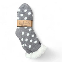 A pair of gray knit home socks with white polka dots and a white sherpa lining at the cuff, packaged with a brown paper band that reads 'Jess & Lou Cozy Homesocks'.