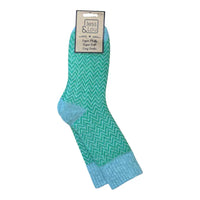 A pair of Jess & Lou crew socks with a green chevron pattern, light green toe and heel sections, and a light green ribbed cuff, hanging from their original paper packaging on a white background.