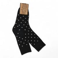 A pair of black crew socks with small white polka dots, hanging from a brown paper brand tag that reads 'Jess & Lou'.