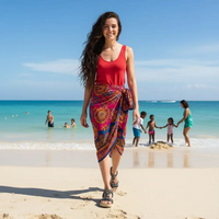 A woman walking on a sandy beach wearing a bright red tank top and a colorful patterned sarong skirt. The background shows a tropical beach scene with turquoise water, blue sky, and other beachgoers.