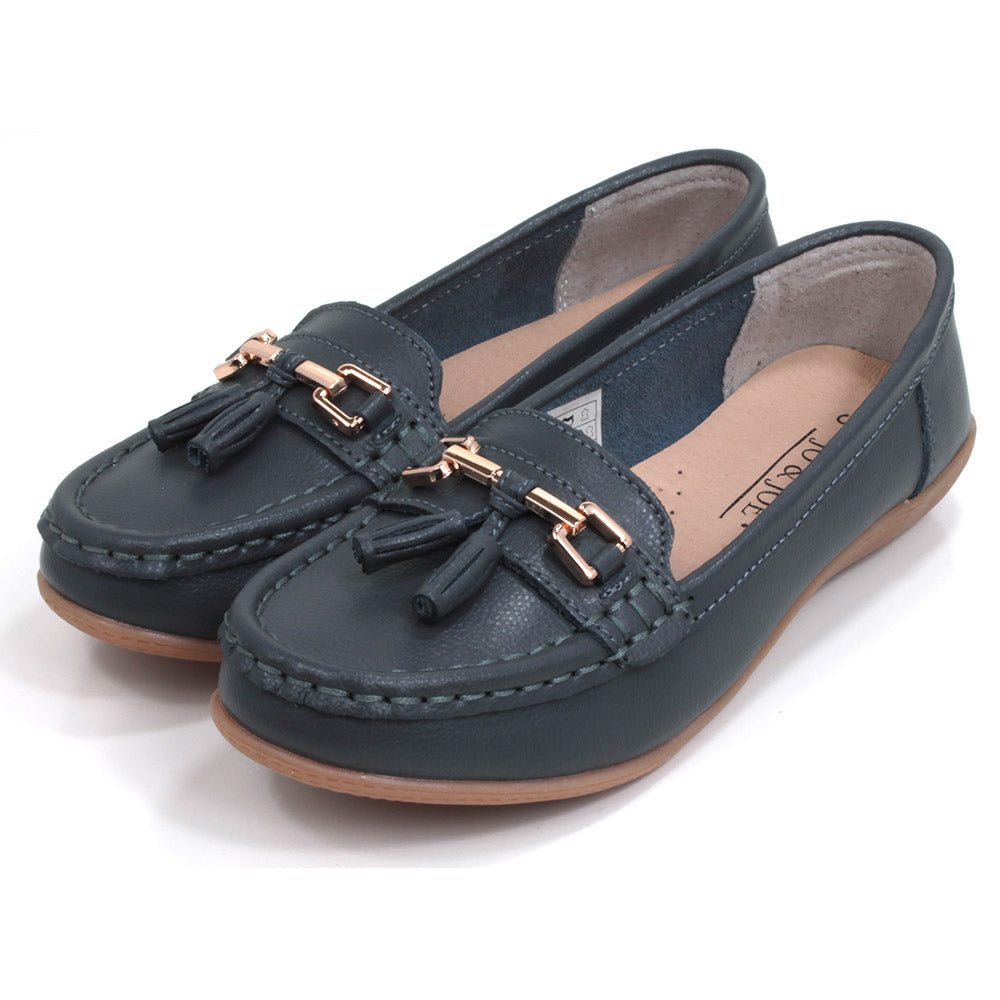 Joe and Jo moccasin style slip on, leather shoes in slate colour. Over the foot metallic gold coloured  detail and two matching tassels. Beige rubber soles. Angled view.