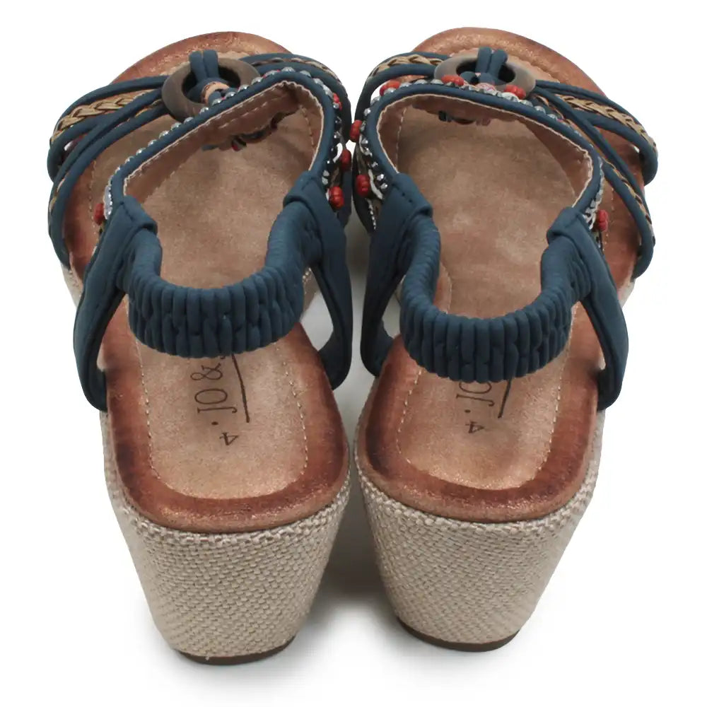 A pair of women's wedge sandals shown from the back, featuring navy blue elastic straps, braided woven straps with small red and white bead accents, a brown leather footbed, and a beige jute-wrapped wedge heel.